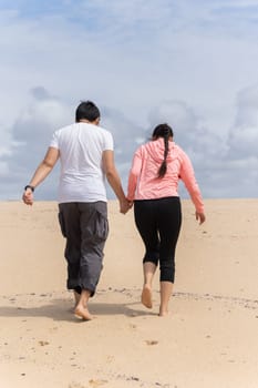 A couple is walking on a sandy beach, holding hands. The woman is wearing a pink hoodie