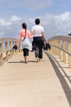 Two people walking on a wooden bridge. One of them is wearing a pink shirt. They are holding their shoes