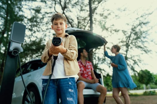 Little boy holding EV charger and point at camera with his family sitting on the trunk in background. Road trip travel with alternative energy charging station for eco-friendly car concept. Perpetual