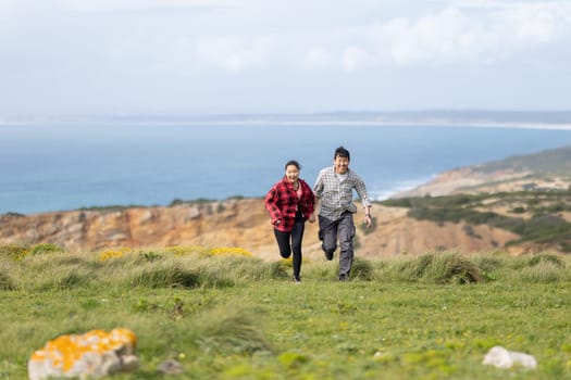A couple running on a grassy hill overlooking the ocean. The man is wearing a plaid shirt and the woman is wearing a red shirt