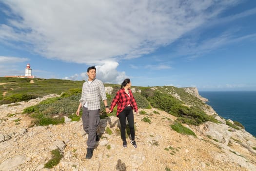 A couple is walking on a rocky hillside with a lighthouse in the background. Scene is peaceful and romantic