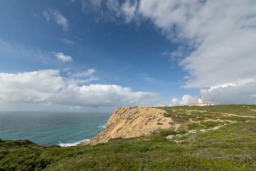 A lighthouse is on a rocky cliff overlooking the ocean. The sky is clear and blue, with a few clouds scattered throughout. The scene is peaceful and serene