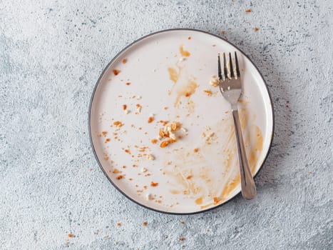 Empty dirty dish after cheesecake with caramel sauce and dessert fork. White rustic trendy modern fashionable dirty plate on gray cement background, top view or flat lay. Copy space for text