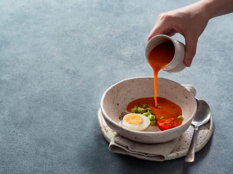 Gaspacho soup and ingredients. Woman's hand pouring of traditional spanish cold soup puree gazpacho in bowl on gray background. Copy space for text or design.