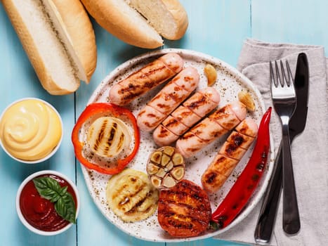 Top view of chicken homemade sausages, buns bread, sauces ketchup and mustard on blue wooden background. Grilled sausages and grilled vegetables in craft trendy plate. Homemade hotdogs.