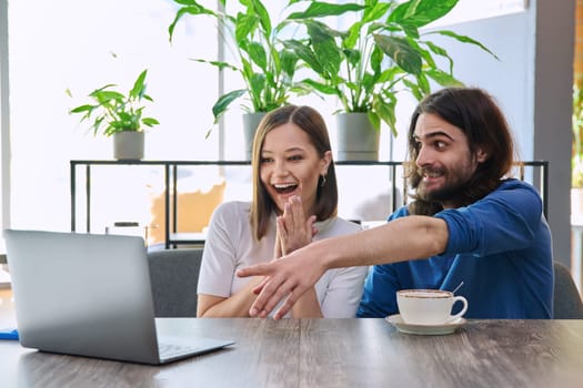 Happy smiling surprised young couple looking at laptop together while sitting in cafe, cafeteria. Leisure time for two, lifestyle, togetherness, relationship, communication, work study remotely