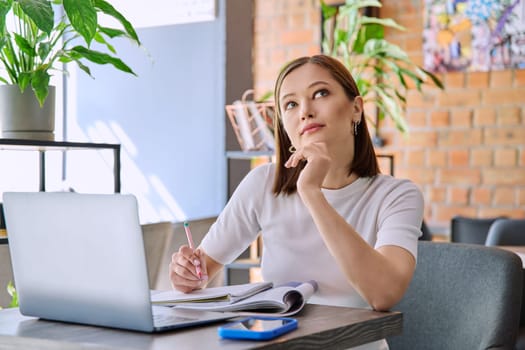 Young female college student studying using laptop, writing in notebook while sitting in coworking cafe. E-learning, education, lifestyle, youth concept