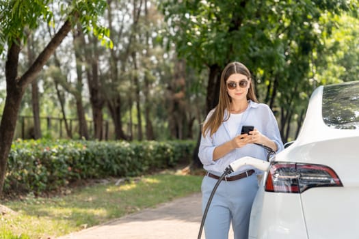 Young woman using smartphone online banking application to pay for electric car battery charging from EV charging station during vacation holiday road trip at national park or summer forest. Exalt