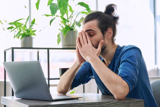 Young tired man working with laptop computer having headache migraine fatigue, holding hands on face. Depression, stress, problems, difficulties with business work, mental health