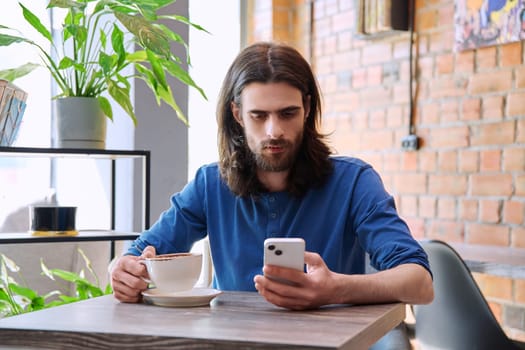 Young 30s stylish handsome bearded with long hair man using smartphone, drinking cup of coffee, sitting in cafe. Mobile technologies Internet apps applications for leisure work business communication