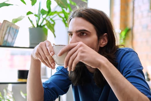 Close up of young handsome man holding drinking cup of coffee while sitting in coffee shop restaurant. Lifestyle, leisure, relaxation, coffee drinks, people concept