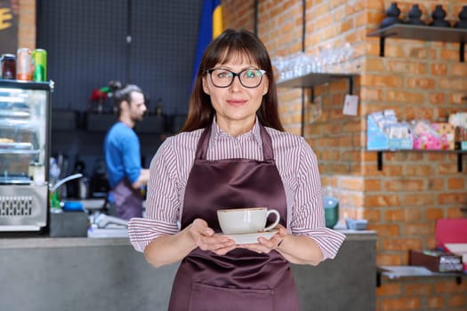 Woman in apron, food service coffee shop worker, small business owner with cup of fresh coffee, looking at camera near bar counter with male barista. Staff, occupation, entrepreneur, work concept