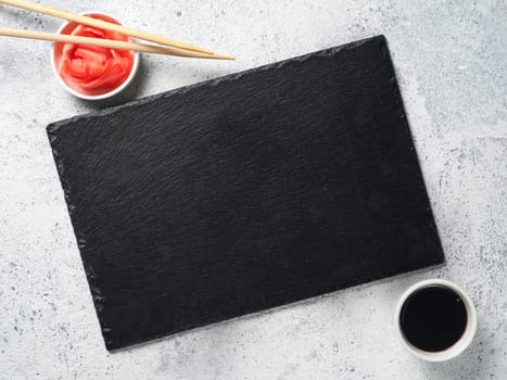 Empty slate plate on gray cement background with chopsticks, pickled ginger and soy sauce. Copy space. Top view or flat-lay. Concept for restaurant and delivery sushi and rolls