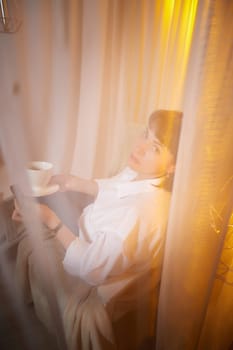 Nice girl with a mug of tea or coffee in cozy room. A middle-aged adult woman relaxing in the evening in the living room or bedroom in the evening