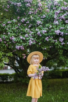 A little girl in a yellow dress and straw hat wearing a bouquet of lilacs. A walk in a spring park, blossoming lilacs