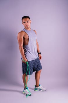 A man with a grey tank top is holding a green jump rope with his elbow bent and waist twisted, showing off his toned chest and wrist strength