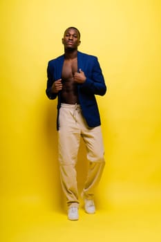A shirtless man in a blue blazer and white pants is posing in front of a yellow wall, showcasing a bold fashion design. His gesture exudes confidence and happiness, perfect for a formal event