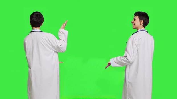 Physician pointing to the left or right to create web commercial standing against greenscreen backdrop, doing a marketing advertisement in studio. Woman medic in white coat shows an ad. Camera B.