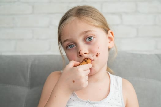 A cute little girl covered in chocolate eats cookies while sitting on the sofa