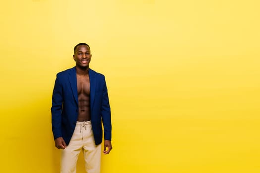 A shirtless man in a blue blazer and white pants is posing in front of a yellow wall, showcasing a bold fashion design. His gesture exudes confidence and happiness, perfect for a formal event