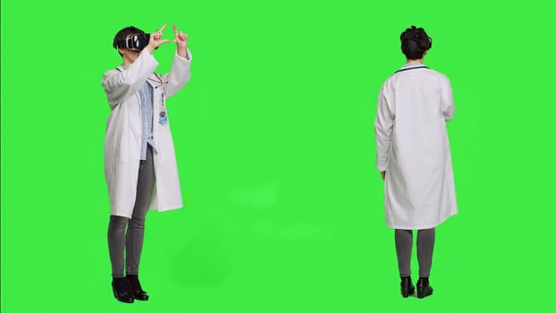 Woman medic works with 3d virtual reality headset in studio, standing against greenscreen backdrop. General practitioner uses vr glasses with interactive vision tool, futuristic exam. Camera A.