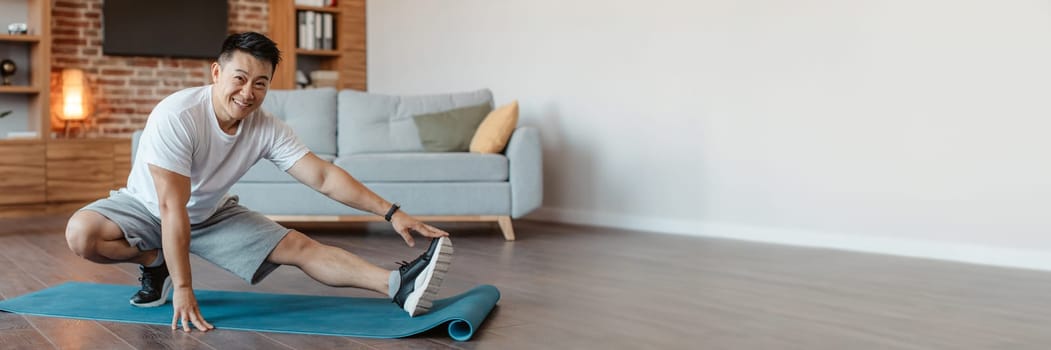 Happy asian mature man training at home, stretching legs in living room interior, panorama with free space. Positive middle aged guy exercising at home, enjoying healthy lifestyle