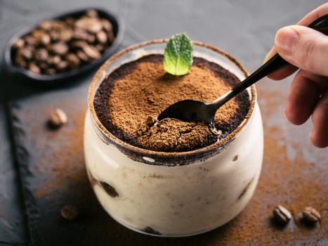 High angle view of tiramisu in glass cup on stone plate over black concrete background, Hand with teaspoon eats tiramisu. Copy space.
