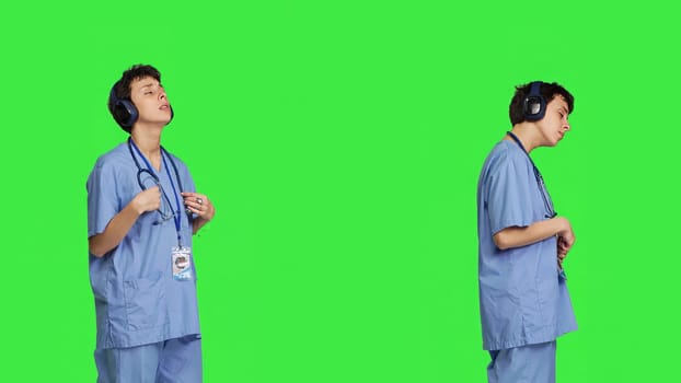 Joyful nurse singing and dancing with wireless headphones on, listening to modern songs against greenscreen backdrop. Specialist enjoying favorite music and creating dance moves. Camera B.