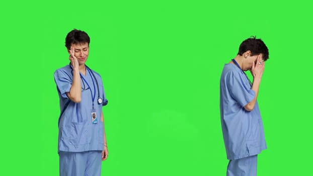 Unwell nurse suffering from a headache against greenscreen backdrop, wearing scrubs and feeling overworked at hospital. Medical assistant having a painful migraine, stress and burnout. Camera B.