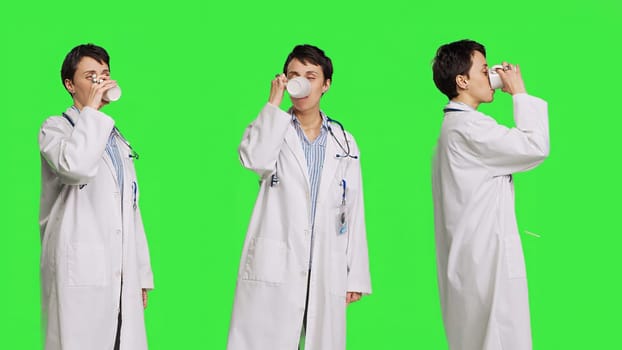 Woman physician drinking a cup of coffee against greenscreen backdrop, taking a break from medical work. Specialist serving caffeine refreshment in studio, wearing a white coat. Camera B.