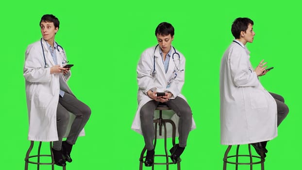 Woman specialist using smartphone social media apps in studio, sitting on a chair against greenscreen backdrop. Physician waiting for someone and browses online webpages, texting. Camera A.