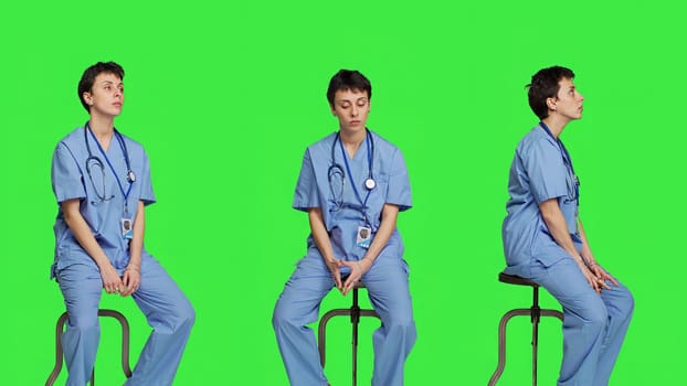 Medical assistant waiting for someone while she sits on chair, feeling impatient and waits for patients to attend checkup appointments. Nurse in scrubs against greenscreen backdrop. Camera A.