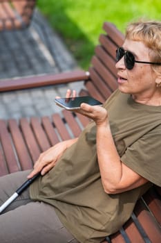 An elderly blind woman sits on a bench in the park and talks on a smartphone. Vertical photo