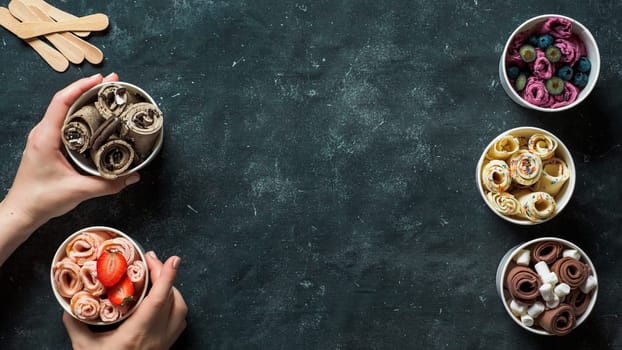 Hands with rolled ice cream in cone cups on dark background. Different iced rolls top view or flat lay. Thai style rolled ice cream with copy space in center for text or design. Banner