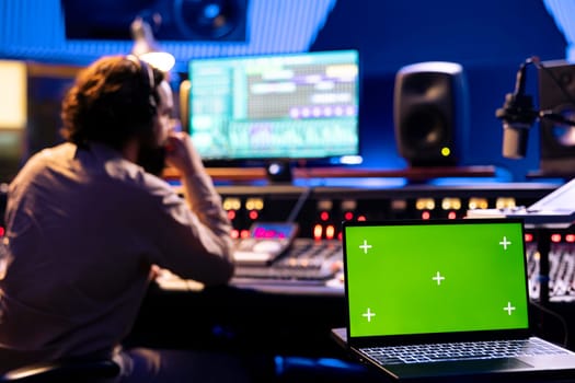 Male technician working on editing tracks next to greenscreen on laptop, operates buttons and knobs in control room. Audio engineer using faders and other technical equipment, mix and master.