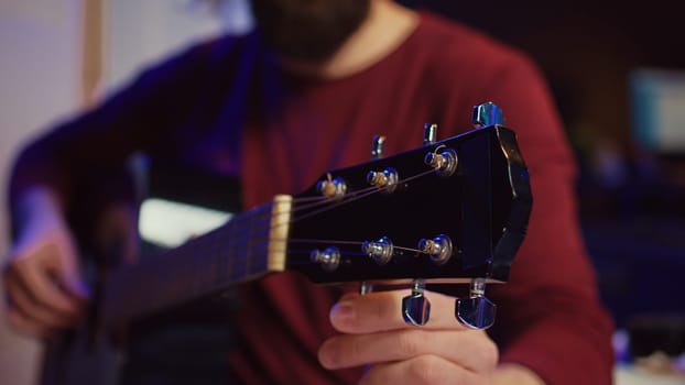 Musical performer tuning his guitar by twisting the knobs, preparing to play acoustic instrument in home studio. Artist recording sounds to create new track, using equalizer mixing gear. Camera B.