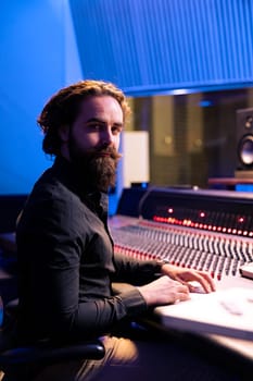 Portrait of sound engineer working with audio console and digital software on pc in professional recording studio, producing tracks for an album. Technician editing new songs in control room.