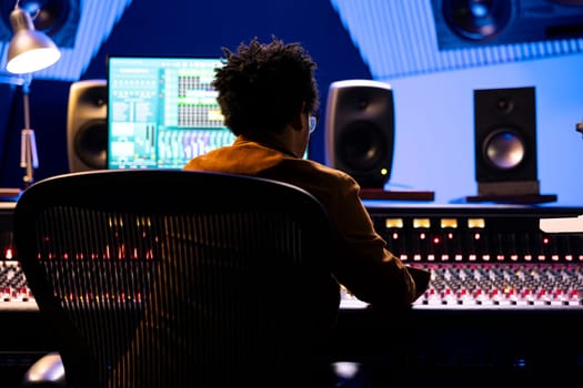 African american record engineer editing and mixing songs on studio console, using digital software on monitor to add audio effects in post production. Producer working on new music.