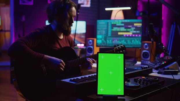Composer performing acoustical guitar in his home studio, playing and practicing on strings. Musician learning multiple songs on instrument, uses tablet to run greenscreen display. Camera B.