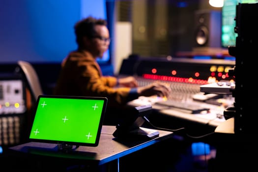 African american sound engineer editing music next to greenscreen display on gadget, operating on control room mixing console. Young producer pressing knobs and sliders in studio.