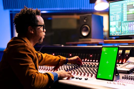 African american music producer recording and editing tracks in studio, operates panel board next to mockup on device. Sound designer mixing and mastering tunes on audio console, stereo gear.