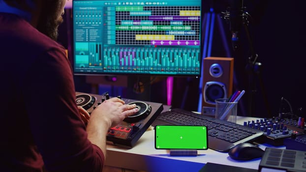 Musical performer creating mixed sounds and tunes with mixing console, greenscreen running on phone display. Artist recording new notes using equalizer and daw software on computer. Camera B.