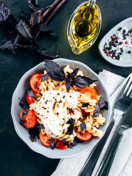 Tasty italian fusilli pasta with cherry, mozarella or buratta cheese and fresh basil. Dish with pasta on black concrete background. Top view. Copy space. Healthy food concept and recipe idea. Vertical