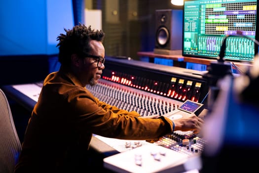 African american music producer composing tracks in control room, editing songs and adjusting volume settings with dashboard knobs. Audio engineer pressing faders and buttons.