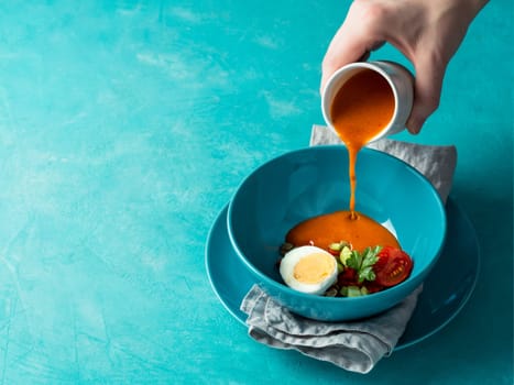 Gaspacho soup and ingredients. Woman's hand pouring of traditional spanish cold soup puree gazpacho in bowl on bright blue background. Copy space for text or design.