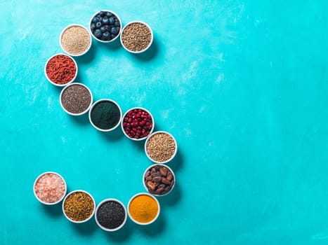 Above view of various superfoods in smal bowl in form S letter on blue background. Superfood as chia,spirulina,cocoa bean,goji, hemp, blueberry, quinoa, bee pollen,black sesame,turmeric. Top view
