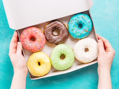 Top view of female hands holding box with colorful donuts on blue concrete background