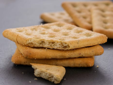 stack of square crackers with pieces and crumbs on slate gray background. Dry salt cracker cookies with fiber and dry spices