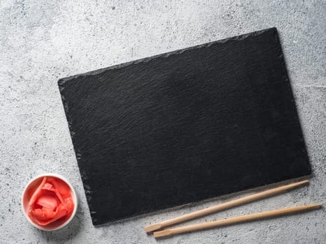 Empty slate plate on gray cement background with chopsticks and pickled ginger. Copy space. Top view or flat-lay. Concept for restaurant and delivery sushi and rolls