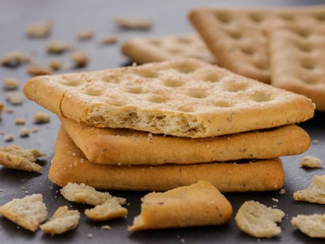 stack of square crackers with pieces and crumbs on slate gray background. Dry salt cracker cookies with fiber and dry spices